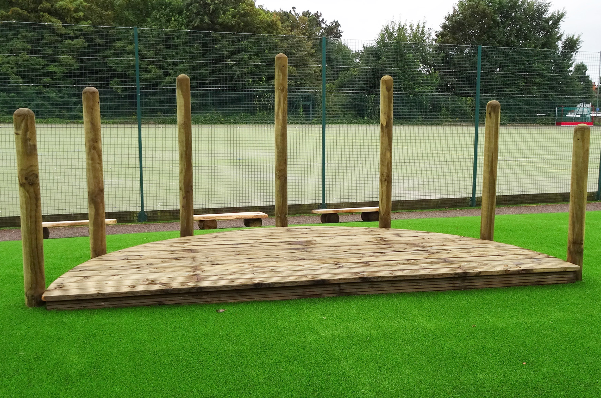 Semi Circular timber stage construction made of decking grooved timber and vertical surround poles sits on green artificial grass