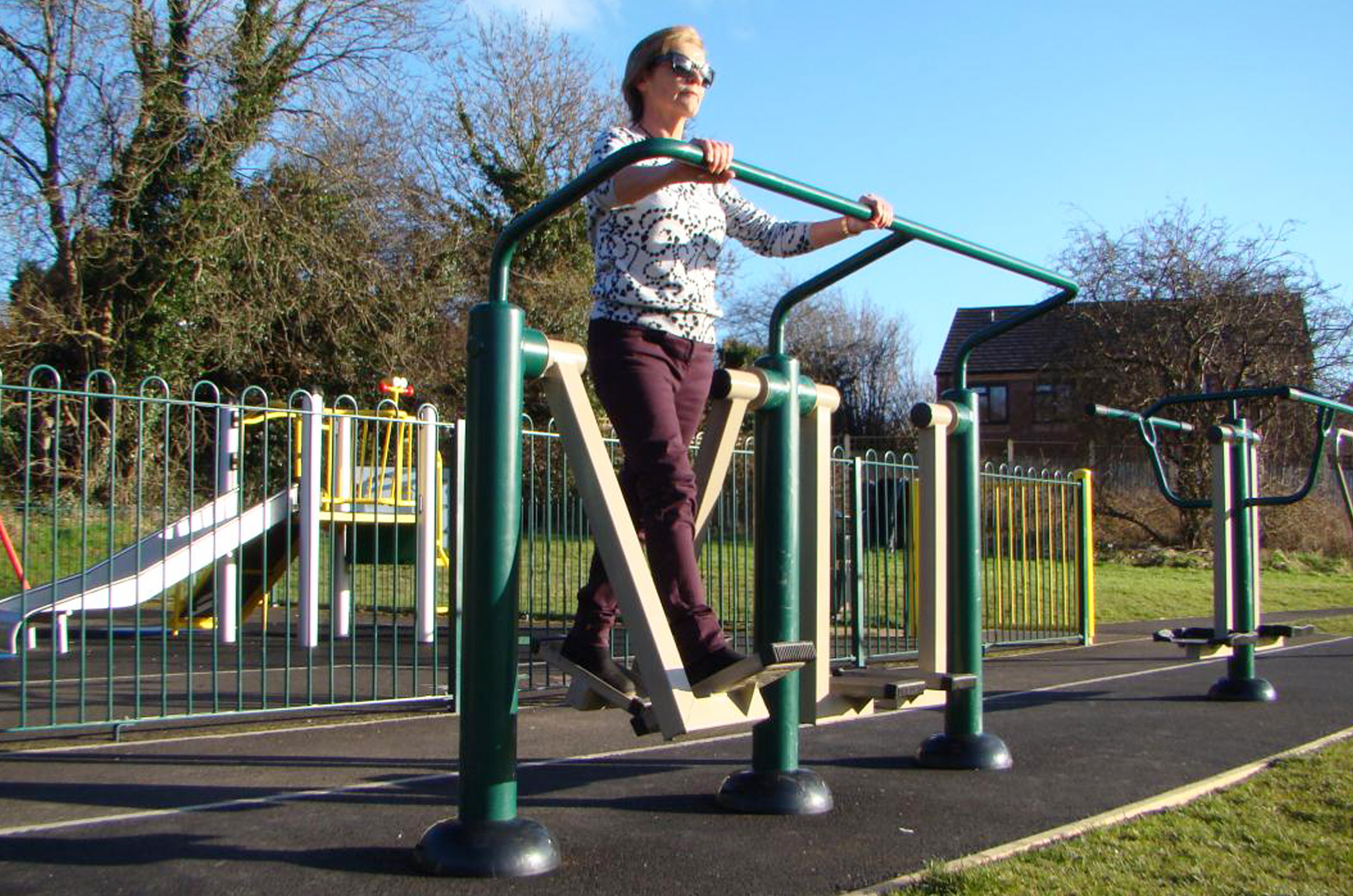 A lady uses one side of the double health walker, her legs are astride in the motion of walking whilst holding on to the support hand rail, the walking motion helps with flexibility in her joints