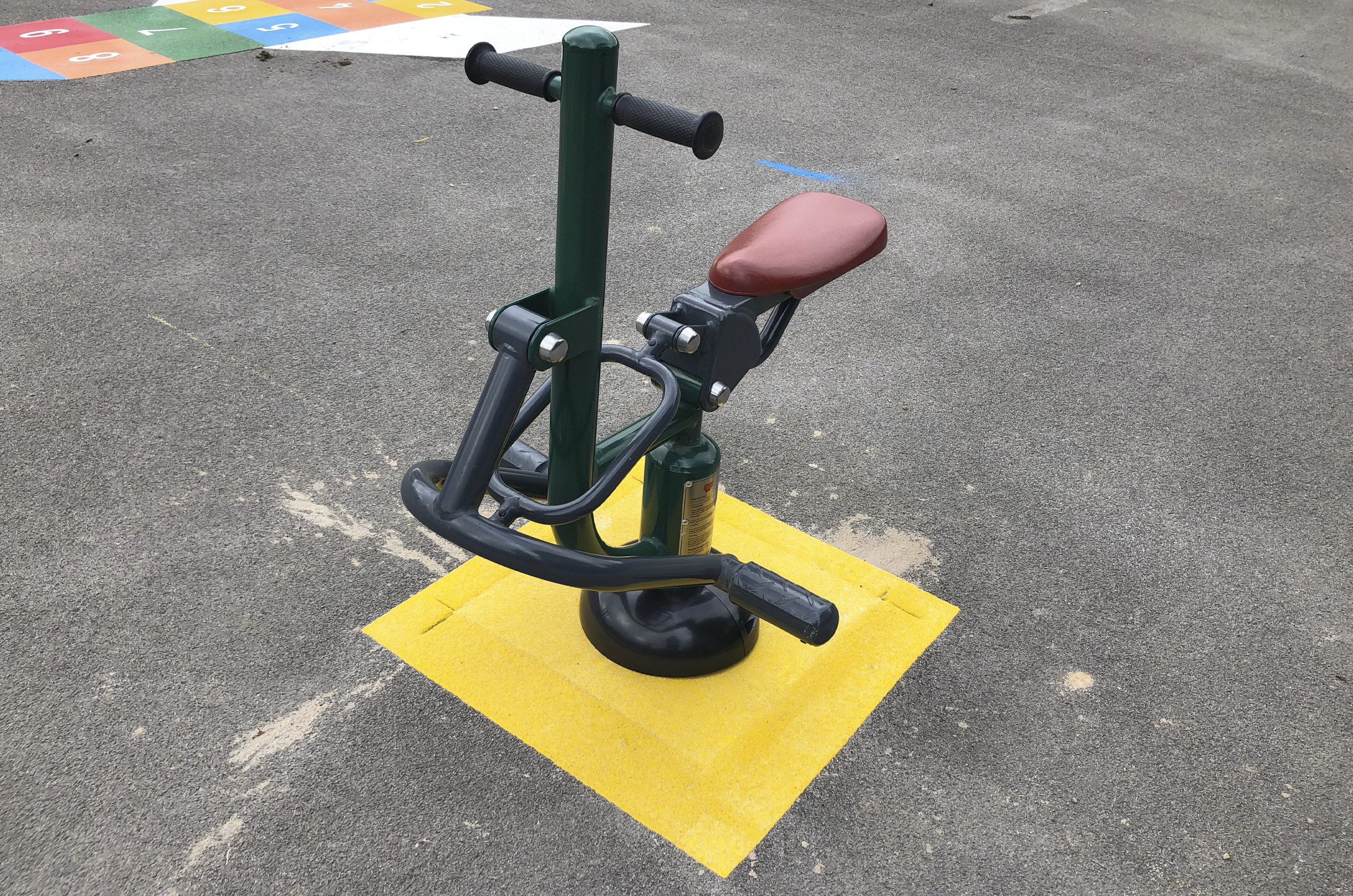 the children's horse rider simulates the up and down motion of riding a horse, it has a seat, two foot holds and a shoulder height handle. it is sitting on a tarmac playground with a yellow coloured square