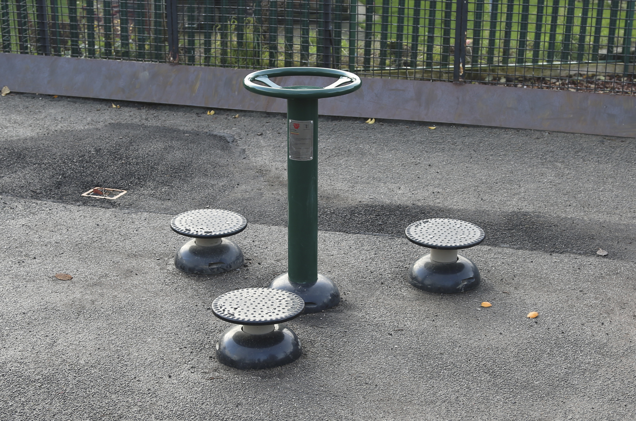 A central green pole with top handles is surrounded by 3 circular discs all sat on tarmac