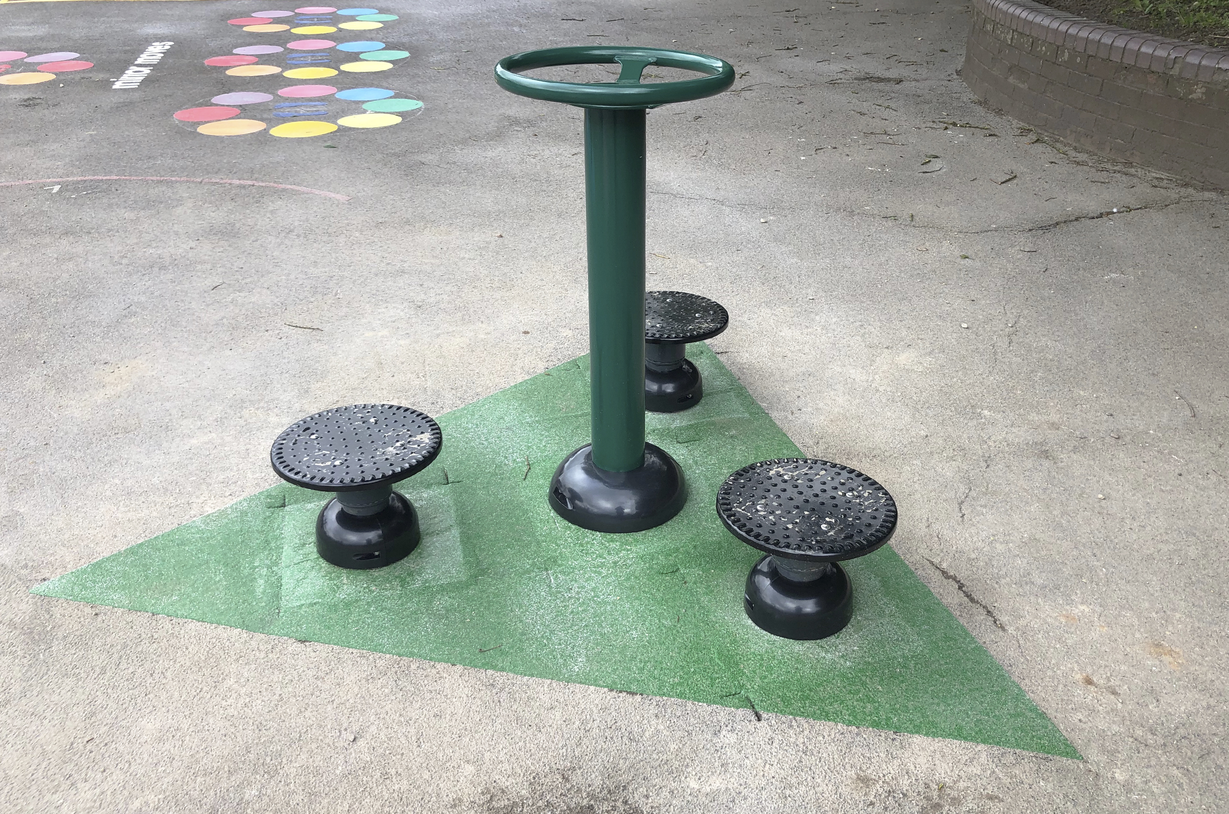 the central post has a waist height circular handle, three outer ground level disc provide a place to stand and rate to twist the waist whilst holding the handle. located on a tarmac playground with a green coloured triangle