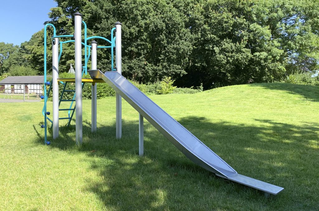 a slide comes down from a platform with rear steps sitting on grass with trees in the background