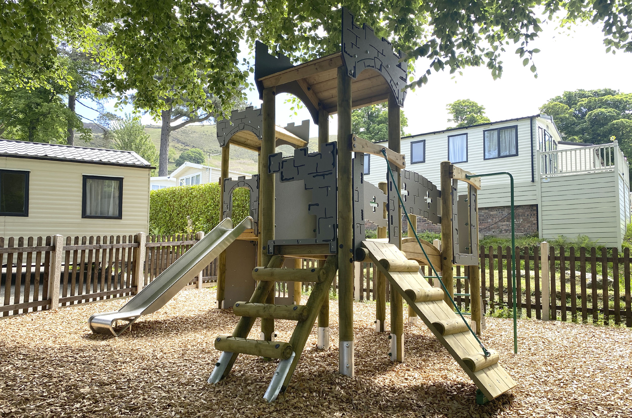 Craiglwyd Hall Caravan Park, a grey castle themed with timber legs play tower with a slide to the left, a ladder an ramp in the middle and a bridge to tower and fire mans pole on the right it sits on wood chip safer surfacing. with trees, a fence and two caravans in the background