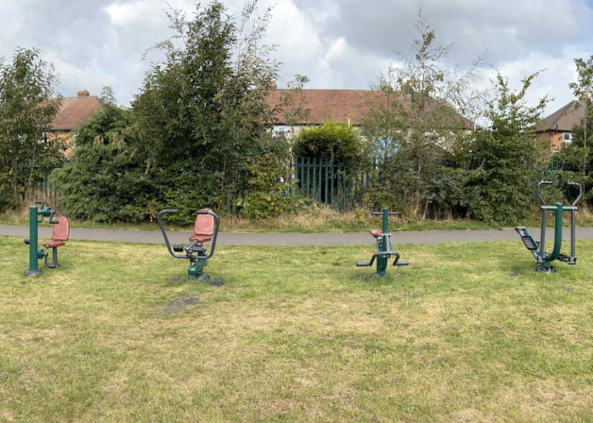 Market Drayton Junior School - Children’s Outdoor Fitness Equipment, seven items of green and grey equipment sit on the grass filed with trees and houses in the background.