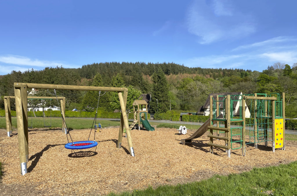 Llanwddyn Community Play Area at Lake Vyrnwy, a timber framed blue and red basket seat swing on the left, a toddler slide in the middle, with a sheep shaped spring seesaw, on the right in a timber climbing frame with steel slide, green nets and yellow climb wall. wood chip safer surfacing with trees and hills with blue sky's in the background.