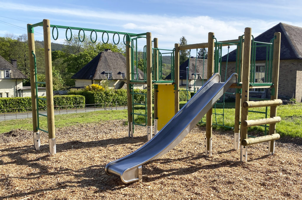 Llanwddyn Community Play Area at Lake Vyrnwy, a timber framed climbing frame, with round Tarzan rings, steel slide, green nets and a log ladder on the right. wood chip safer surfacing, with houses and blue sky's in the background.