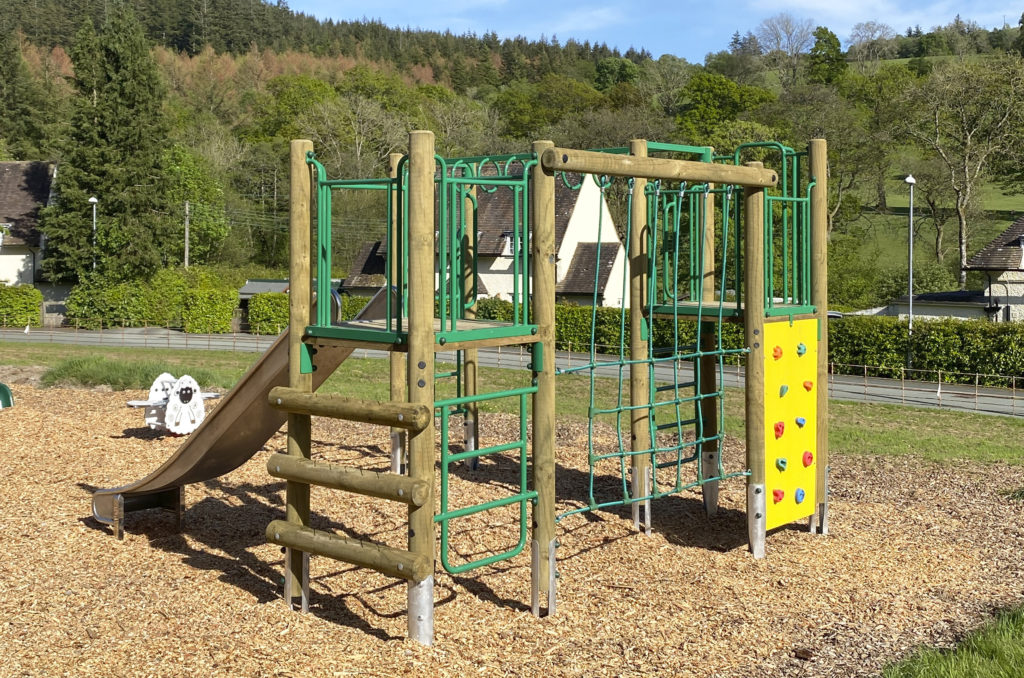 Llanwddyn Community Play Area at Lake Vyrnwy, a sheep shaped spring seesaw on the left, on the right in a timber climbing frame with steel slide, green nets and yellow climb wall. wood chip safer surfacing with houses, trees and hills with blue sky's in the background.