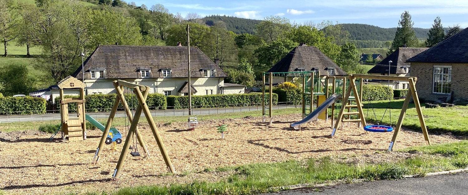 Llanwddyn Community Play Area at Lake Vyrnwy, a toddler slide in and timber framed flat swings on the left, with a sheep shaped spring seesaw and curlicue green spinner in the middle, on the right in a timber climbing frame with steel slide, green nets and yellow climb wall and a timber frame swing with blue and red basket seat. wood chip safer surfacing, with houses hills and blue sky's in the background.