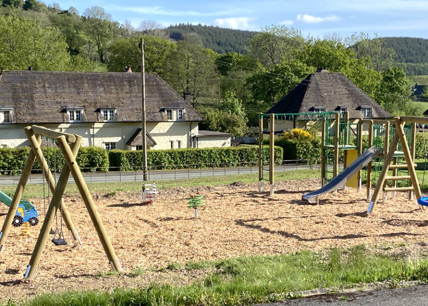 Llanwddyn Community Play Area at Lake Vyrnwy, a toddler slide in and timber framed flat swings on the left, with a sheep shaped spring seesaw and curlicue green spinner in the middle, on the right in a timber climbing frame with steel slide, green nets and yellow climb wall and a timber frame swing with blue and red basket seat. wood chip safer surfacing, with houses hills and blue sky's in the background.