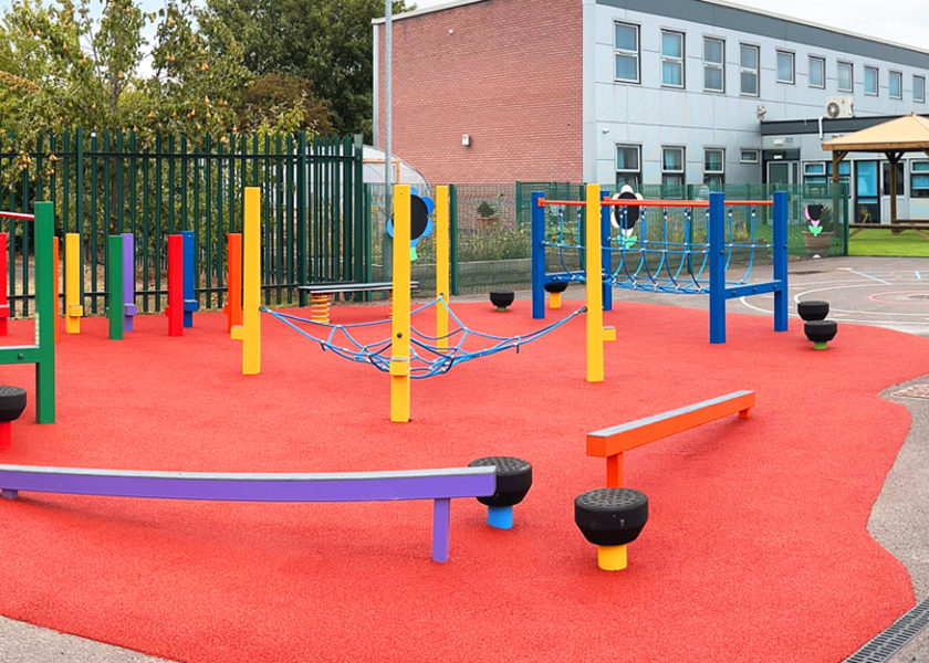 Severndale Specialist Academy. Playground with multicoloured steel play equipment on a red wetpour safer surface. Trees and school buildings in the background.