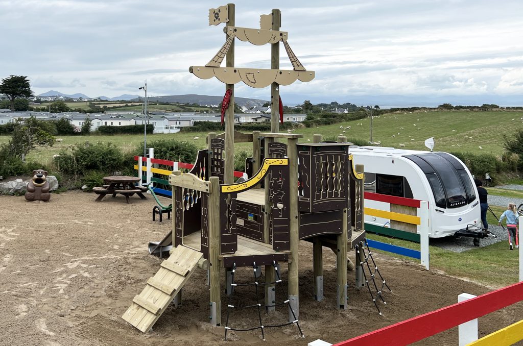 Deucoch Touring and Camping Park Abersoch Crows Nest Multiplay, timber poles with dark brown shipped themed play panels and lighter brown flags and sails, below is sand safer surfacing. in the back ground there is white caravans and green hills.