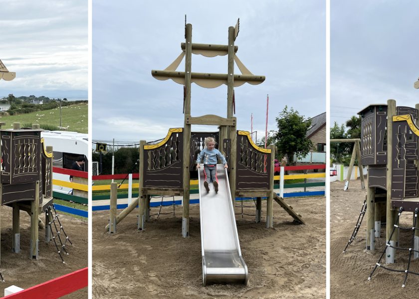 Deucoch Touring and Camping Park Abersoch, timber poles with dark brown shipped themed play panels and lighter brown flags and sails, in the the centre there is a silver slide with a young girl. below is sand safer surfacing.