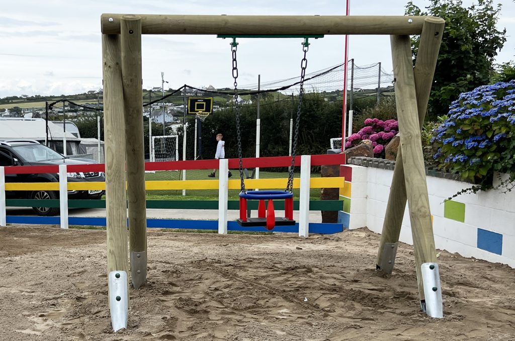 Deucoch Touring and Camping Park Abersoch Cradle Swing, a set of timber poles hold up and red and blue cradle swing seat. below is sand safer surfacing. a colourful fence in in the background.