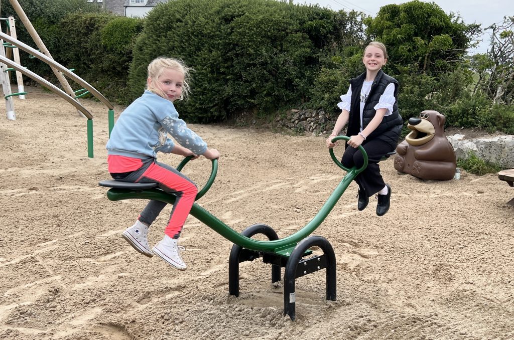 Deucoch Touring and Camping Park Abersoch Seesaw, A green and black frame with green circular handles. with young girls are sat on the seesaw smiling. below is sand safer surfacing. green hedge is in the background.